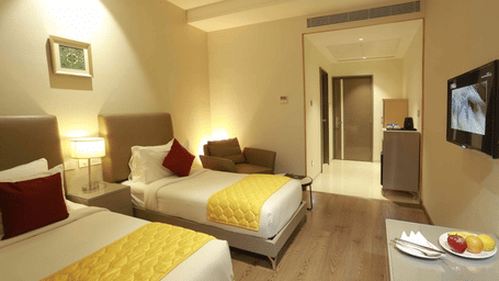 Premium Twin Room at Mayberry Tiruppur featuring a TV stand, wall-mounted TV, and a view of the outside door.